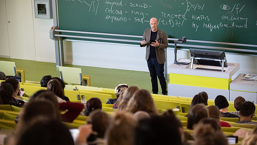 Image: Teaching situation in a lecture hall. The lecturer stands in front of the blackboard and speaks to the students.