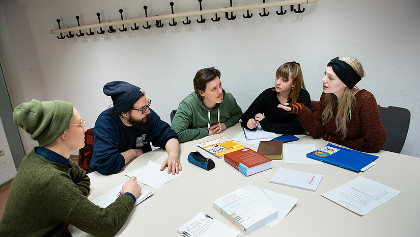 Image: Teaching and working situation in a seminar room. A group of five students are sitting at a table and discussing. There are textbooks and work materials on the table.