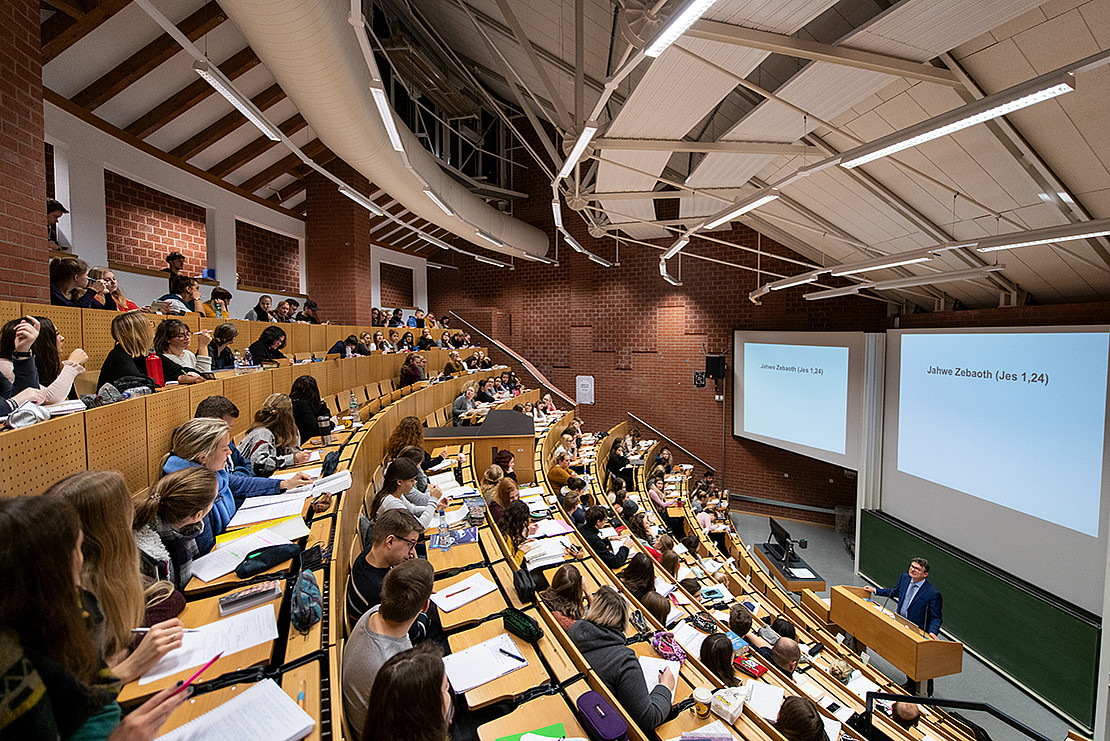 Image of a teaching situation. A small lecture hall can be seen. In the rows of seats, the students have their work materials in front of them and are taking handwritten notes. The lecturer is standing downstairs in front of his lectern, with the slides for the lecture projected onto screens in the background.