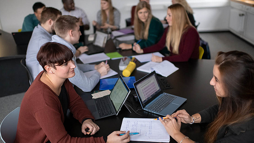 Imagebild: Lern- und Arbeitssituation. Eine Gruppe von elf Studierenden sitzt an Tischen in einem Seminarraum. Auf den Tischen stehen einige Laptops und es sind Arbeitsmaterialien ausgebreitet. In Kleingruppen unterhalten sie sich.