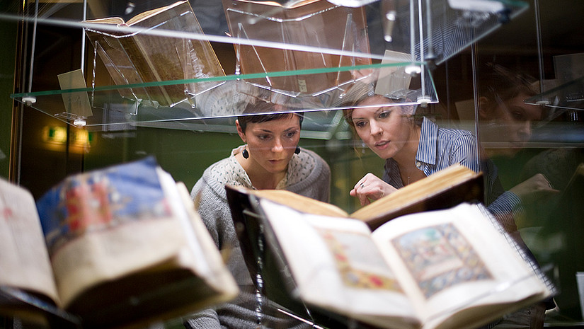 Imagebild: Zwei Personen schauen auf die ausgestellten kunsthistorischen Bücher in einer gläsernen Vitrine. 