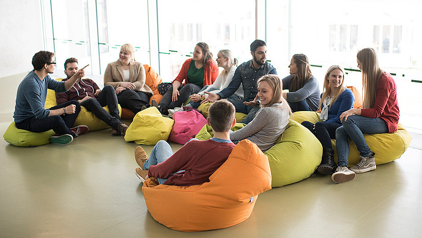 Imagebild einer Lehr- und Arbeitssituation. Eine Gruppe von elf Studierenden befindet sich in einem kleinen Raum. Im Hintergrund sind bodentiefe Fenster zu sehen. Davor sind Sitzsäcke platziert, auf denen die Studierenden sitzen und sich unterhalten.