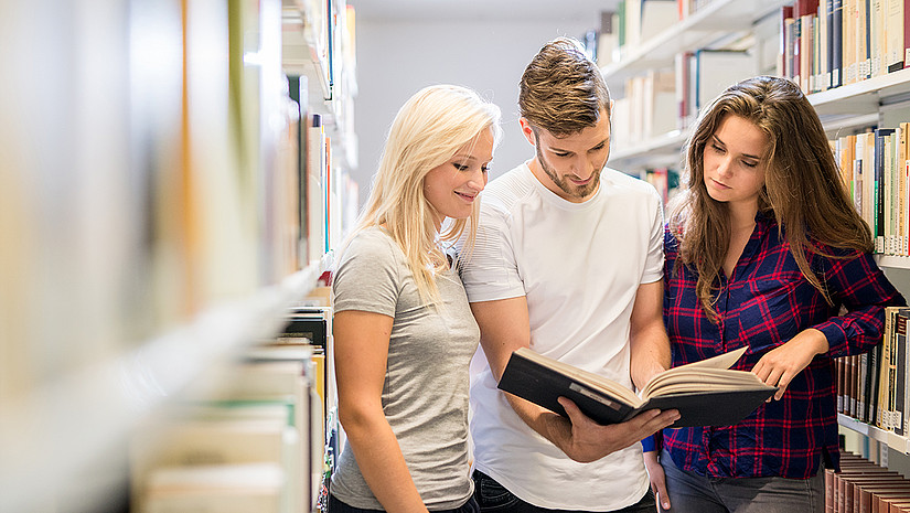 Imagebild: Drei Studierende stehen zwischen den Bücherregalen in der Bibliothek. Einer der Studierenden hält ein geöffnetes Buch in der Hand, welches sie gemeinsam betrachten.