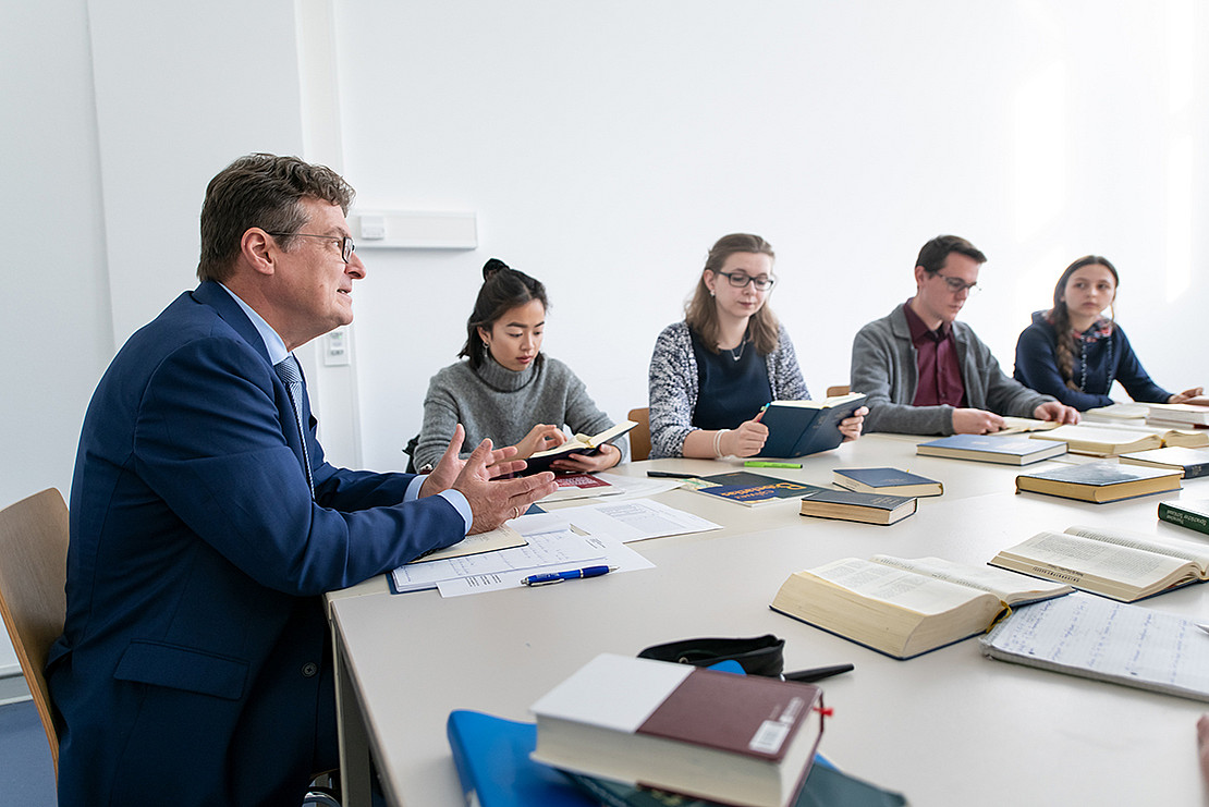 Image of a teaching and working situation. Four students are depicted sitting together with the lecturer at tables arranged in a seminar room. The lecturer is speaking and the students are looking at the books open in front of them. There are other books and work materials on the tables.