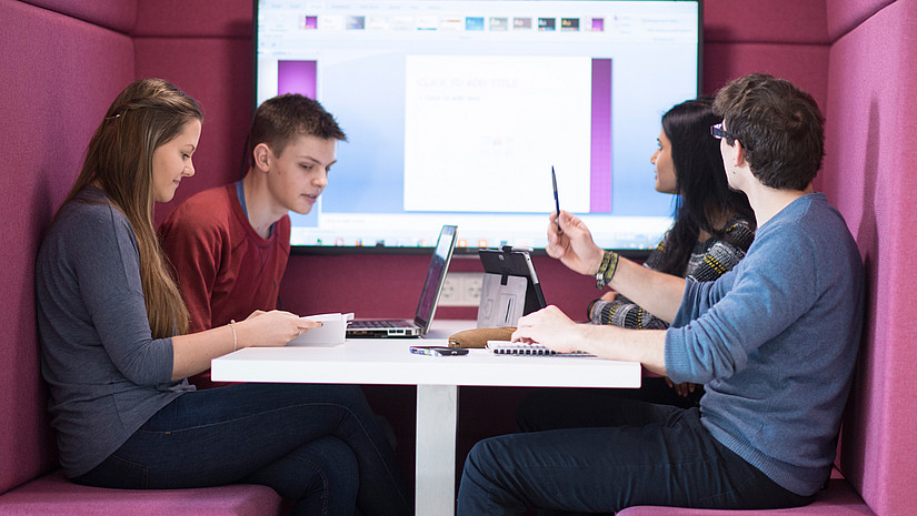 Imagebild: Studierende in einer Arbeitssituation. Vier Studierende sitzen gemeinsam an einem Tisch. Hinter ihnen ist ist ein Präsentationsmonitor zu sehen.